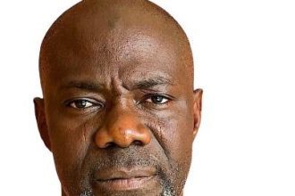 A close-up headshot of a middle-aged Black man with a bald head and a distinct white and grey beard, wearing a green shirt, looking directly at the camera.