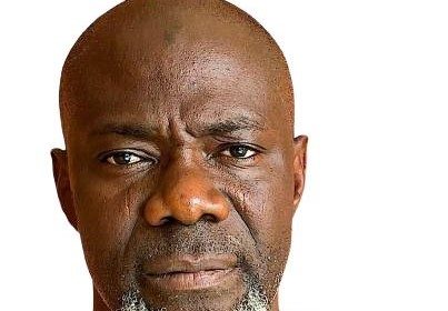 A close-up headshot of a middle-aged Black man with a bald head and a distinct white and grey beard, wearing a green shirt, looking directly at the camera.