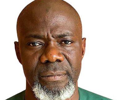 A close-up headshot of a middle-aged Black man with a bald head and a distinct white and grey beard, wearing a green shirt, looking directly at the camera.